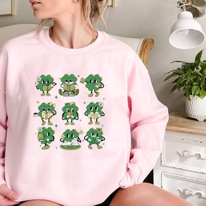 May include: A pink sweatshirt with a graphic of nine green four-leaf clovers holding books and cups. Each clover has a different expression and is holding a different book or cup.