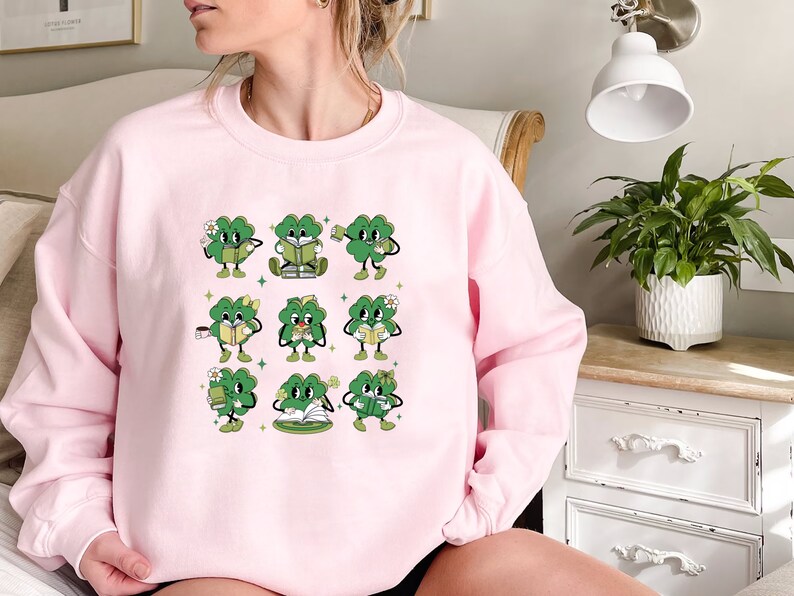 May include: A pink sweatshirt with a graphic of nine green four-leaf clovers holding books and cups. Each clover has a different expression and is holding a different book or cup.