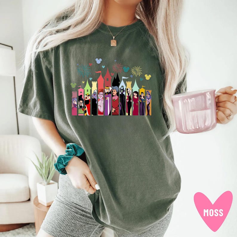 Puede incluir: Camiseta verde musgo con un gr&aacute;fico colorido de villanos de dibujos animados frente a un horizonte de castillo con fuegos artificiales e iconos de Mickey Mouse. La camiseta es de corte holgado.
