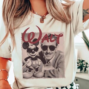 May include: A cream-colored t-shirt featuring a graphic print of Walt Disney and Mickey Mouse both wearing sunglasses. The word "Walt" is printed in a large, dark red script font above the black and white image of the two figures.