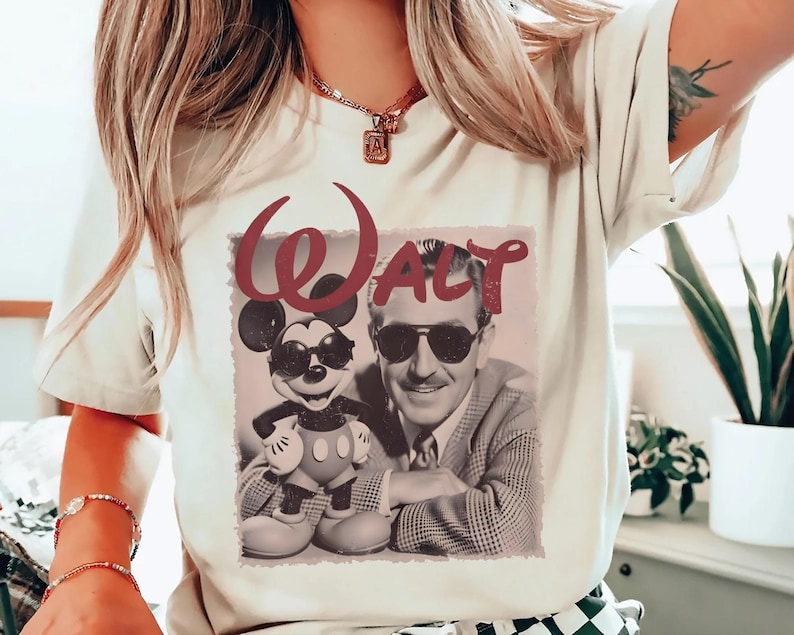 May include: A cream-colored t-shirt featuring a graphic print of Walt Disney and Mickey Mouse both wearing sunglasses. The word "Walt" is printed in a large, dark red script font above the black and white image of the two figures.