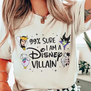 以下が含まれることがあります： 「99% SURE I AM A Disney VILLAIN」の文字とディズニーヴィランズのイラストが描かれたクリーム色のTシャツ。デザインには、邪悪な女王、マレフィセント、アースラ、クルエラが含まれています。Tシャツはクルーネックで半袖です。