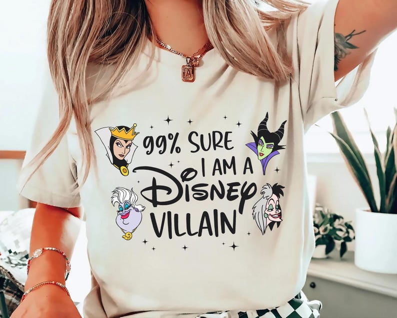 以下が含まれることがあります： 「99% SURE I AM A Disney VILLAIN」の文字とディズニーヴィランズのイラストが描かれたクリーム色のTシャツ。デザインには、邪悪な女王、マレフィセント、アースラ、クルエラが含まれています。Tシャツはクルーネックで半袖です。