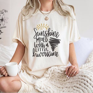 Op de afbeelding: Een wit T-shirt met een zwarte en gouden afbeelding met de tekst "Sunshine mixed with a little hurricane".