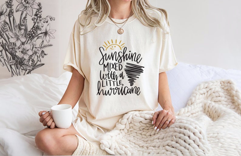 Op de afbeelding: Een wit T-shirt met een zwarte en gouden afbeelding met de tekst "Sunshine mixed with a little hurricane".