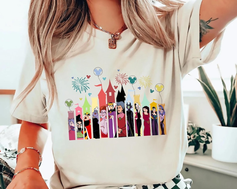 Puede incluir: Camiseta color crema con un gr&aacute;fico colorido de villanos de dibujos animados frente a un castillo. El gr&aacute;fico incluye fuegos artificiales y globos. La camiseta es una prenda casual para el d&iacute;a a d&iacute;a.