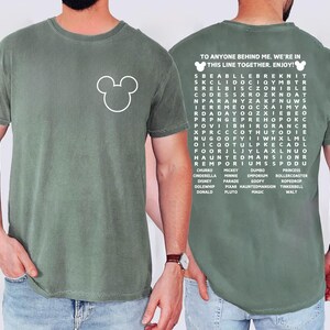 May include: Sage green t-shirt featuring a white Mickey Mouse outline on the front. The back of the shirt has a word search puzzle with the text "TO ANYONE BEHIND ME. WE'RE IN THIS LINE TOGETHER. ENJOY!" and Disney-related words.