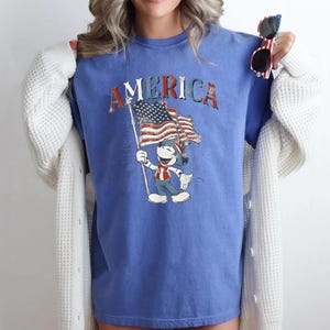 Peut inclure: T-shirt bleu avec le mot "AMERICA" en lettres rouges, blanches et bleues. Le t-shirt pr&eacute;sente un dessin d'un personnage de dessin anim&eacute; tenant un drapeau am&eacute;ricain. Le personnage porte une tenue patriotique.