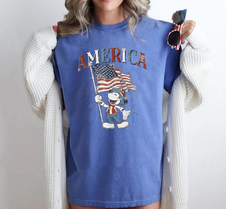 Peut inclure: T-shirt bleu avec le mot "AMERICA" en lettres rouges, blanches et bleues. Le t-shirt pr&eacute;sente un dessin d'un personnage de dessin anim&eacute; tenant un drapeau am&eacute;ricain. Le personnage porte une tenue patriotique.