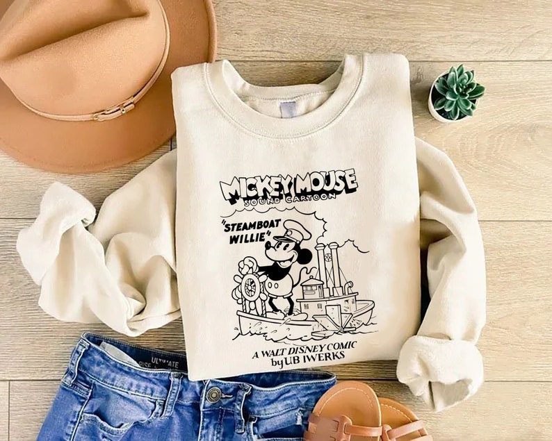 K&ouml;nnte beinhalten: Ein cremefarbener Pullover mit einer Schwarzwei&szlig;illustration von Mickey Mouse auf einem Dampfschiff. Der Text "Mickey Mouse Sound Cartoon" befindet sich &uuml;ber dem Bild und "Steamboat Willie" darunter. Der Text "A Walt Disney Comic by Ub Iwerks" befindet sich am unteren Rand des Bildes.