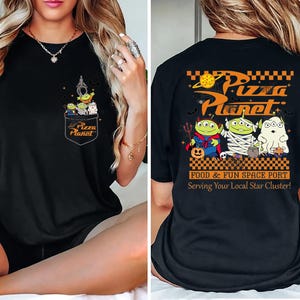 Pode incluir: Camiseta preta com um design de bolso com alien&iacute;genas de desenho animado e o texto "Pizza Planet". A parte de tr&aacute;s da camiseta exibe "Pizza Planet" em laranja, com alien&iacute;genas de desenho animado em fantasias de Halloween. O texto nas costas diz "Food & Fun Space Port."