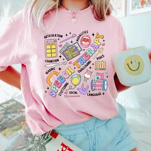 May include: Pink t-shirt with a colorful graphic design that says "Speech Therapy" and includes illustrations of various speech therapy tools and symbols.