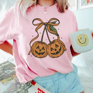 May include: A light pink t-shirt featuring a Halloween-themed graphic. The graphic depicts two orange jack-o'-lanterns with smiling faces, connected by a brown stem and bow. The shirt is paired with light blue shorts.