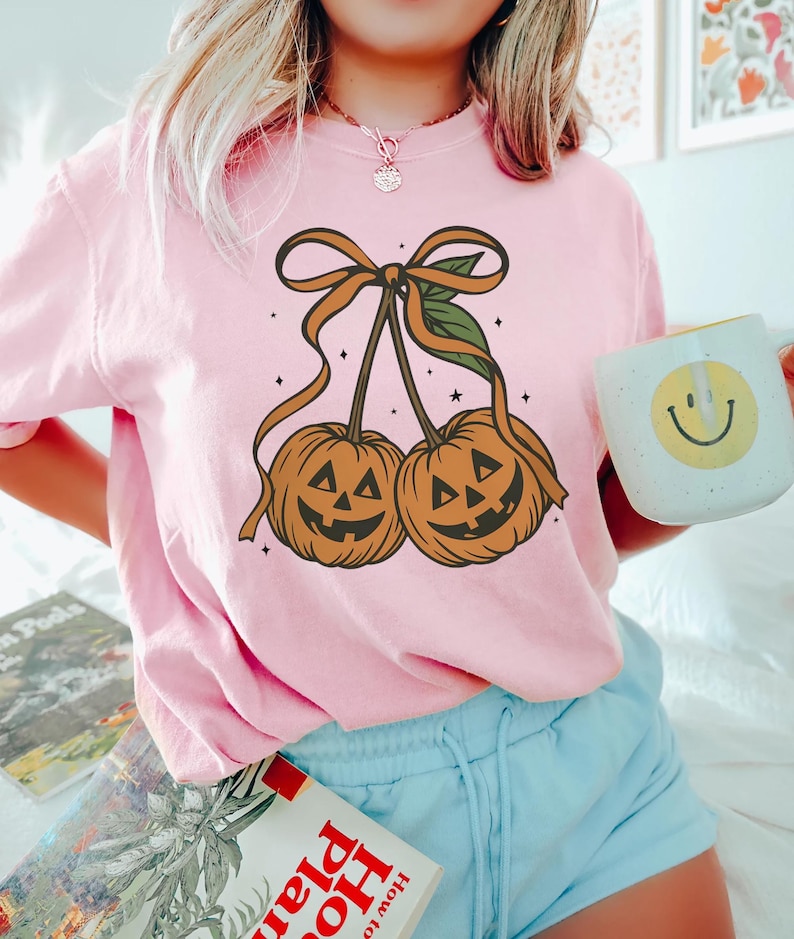 May include: A light pink t-shirt featuring a Halloween-themed graphic. The graphic depicts two orange jack-o'-lanterns with smiling faces, connected by a brown stem and bow. The shirt is paired with light blue shorts.
