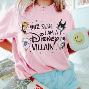 以下が含まれることがあります： 「99% Sure I Am A Disney Villain」の文字とディズニーヴィランのイラストがプリントされた、明るいピンク色のTシャツ。明るいブルーのショートパンツと合わせています。黄色いスマイリーフェイスの白いマグカップが手に持たれています。