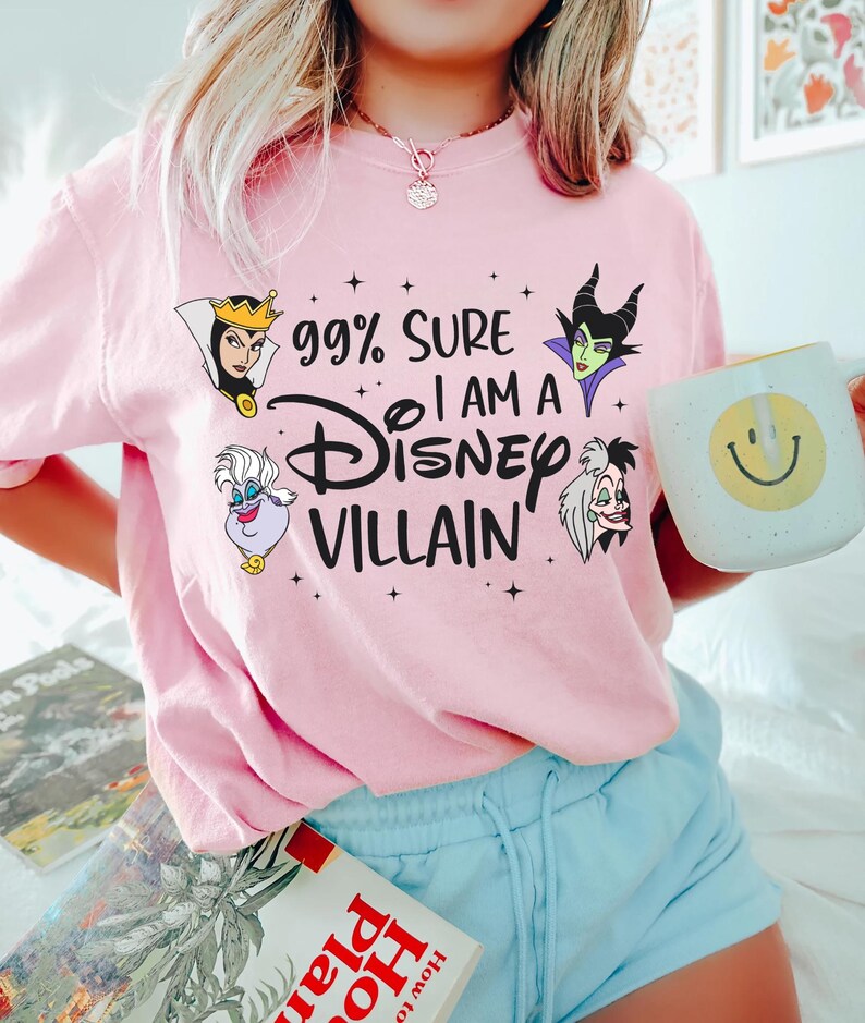 以下が含まれることがあります： 「99% Sure I Am A Disney Villain」の文字とディズニーヴィランのイラストがプリントされた、明るいピンク色のTシャツ。明るいブルーのショートパンツと合わせています。黄色いスマイリーフェイスの白いマグカップが手に持たれています。