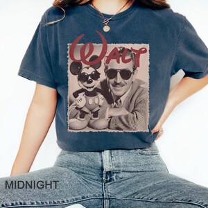 May include: A dark blue short-sleeve t-shirt featuring a graphic print of Walt Disney and Mickey Mouse both wearing sunglasses. The word Walt is printed in large red letters above the figures. The shirt has a crew neckline and a relaxed fit.