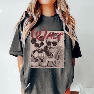 May include: A charcoal gray short-sleeve t-shirt featuring a graphic print of a man and a cartoon mouse both wearing sunglasses. The word WALT is printed in a large, distressed red font above the image, which is set against a light background.