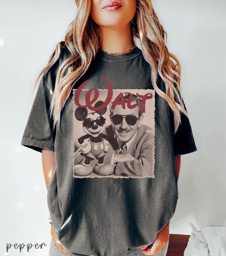May include: A charcoal gray short-sleeve t-shirt featuring a graphic print of a man and a cartoon mouse both wearing sunglasses. The word WALT is printed in a large, distressed red font above the image, which is set against a light background.