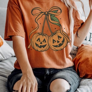 May include: Orange t-shirt featuring a Halloween-themed design. The graphic depicts two jack-o'-lanterns with smiling faces, connected by a bow and ribbon. The shirt is a solid orange color, perfect for the fall season.
