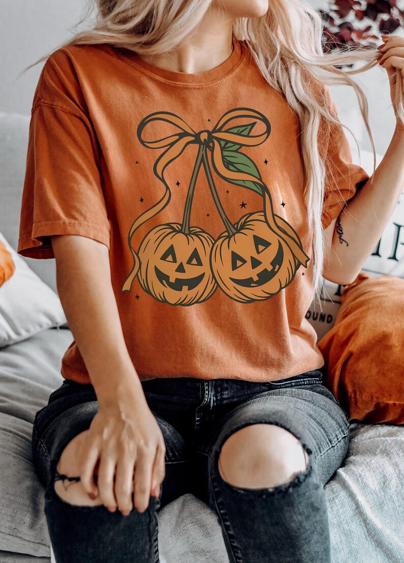 May include: Orange t-shirt featuring a Halloween-themed design. The graphic depicts two jack-o'-lanterns with smiling faces, connected by a bow and ribbon. The shirt is a solid orange color, perfect for the fall season.