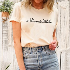 May include: A cream colored t-shirt with the text "Alhamdulillah" written in black cursive script.