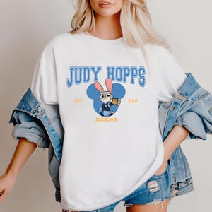 Op de afbeelding: Wit T-shirt met een blauwe afbeelding van Judy Hopps uit Zootopia. Het ontwerp bevat de tekst "JUDY HOPPS" in blauwe blokletters, een cartoonafbeelding van Judy Hopps en de tekst "EST. 2016" en "ZOOTOPIA".