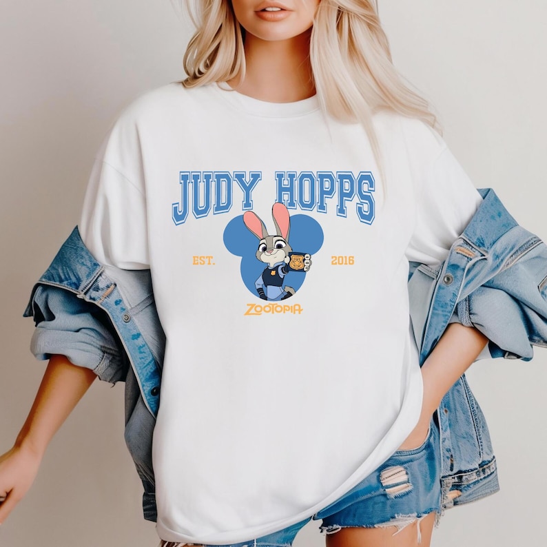 Op de afbeelding: Wit T-shirt met een blauwe afbeelding van Judy Hopps uit Zootopia. Het ontwerp bevat de tekst "JUDY HOPPS" in blauwe blokletters, een cartoonafbeelding van Judy Hopps en de tekst "EST. 2016" en "ZOOTOPIA".