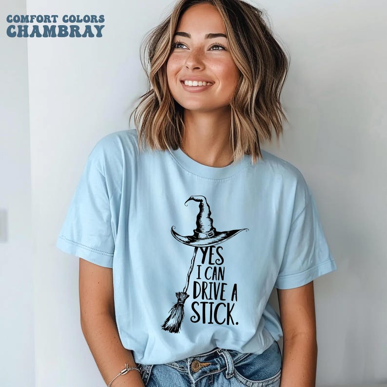 以下が含まれることがあります： 「YES I CAN DRIVE A STICK」の文字が入った水色のTシャツ。テキストの下には、黒い魔女の帽子とほうきのグラフィックが描かれています。シャツの上部には「COMFORT COLORS CHAMBRAY」の文字もプリントされています。
