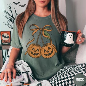 May include: A moss green t-shirt featuring a graphic of two jack-o'-lanterns with orange ribbon and leaves. A black mug with a white ghost design is held in the hand. The word "MOSS" is printed on the bottom right.