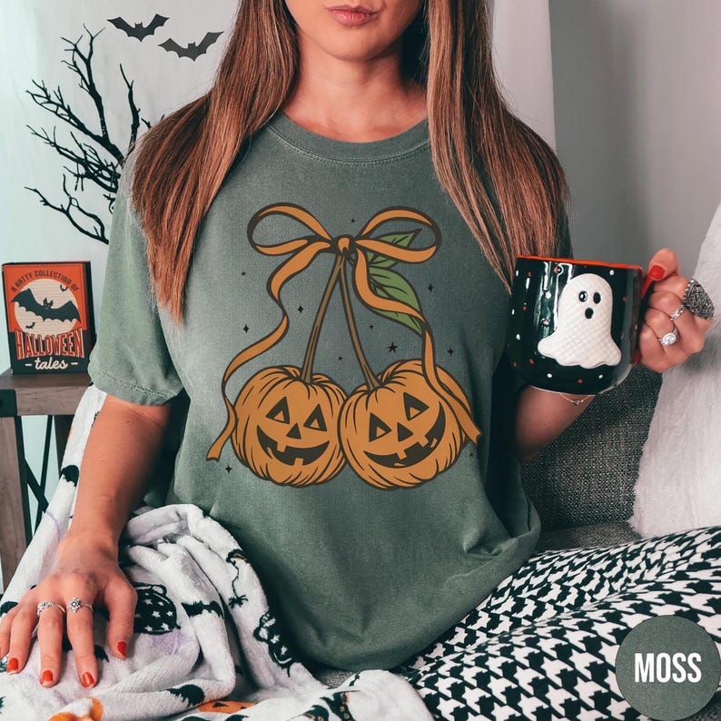 May include: A moss green t-shirt featuring a graphic of two jack-o'-lanterns with orange ribbon and leaves. A black mug with a white ghost design is held in the hand. The word "MOSS" is printed on the bottom right.