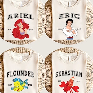 May include: Four white t-shirts with different Disney characters printed on them. The shirts feature Ariel, Eric, Flounder, and Sebastian from The Little Mermaid. Each shirt has the character's name printed in black with the text "EST. 1989" underneath.