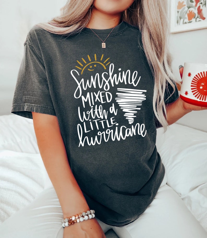 Op de afbeelding: Een donkergrijs T-shirt met een witte afbeelding waarop staat "Sunshine mixed with a little hurricane". De afbeelding bevat een gele zon en een witte tornado.