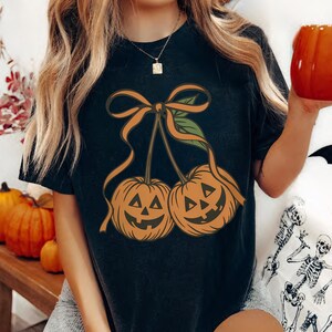 May include: Black t-shirt featuring a Halloween-themed design. The graphic depicts two orange jack-o'-lanterns with smiling faces, connected by an orange bow and green leaves. The shirt is a comfortable, casual style.