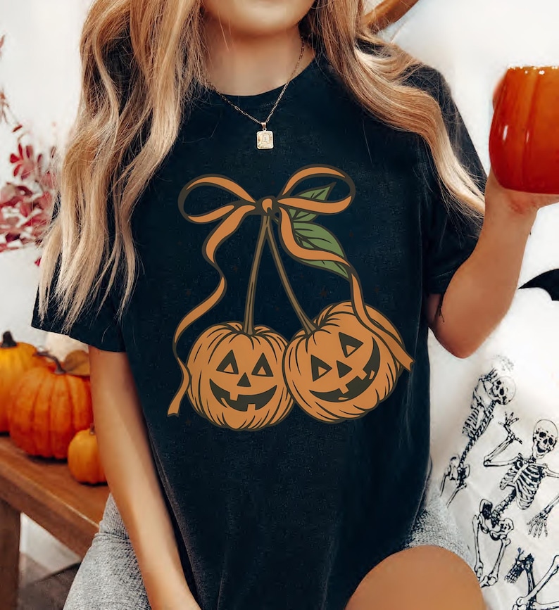 May include: Black t-shirt featuring a Halloween-themed design. The graphic depicts two orange jack-o'-lanterns with smiling faces, connected by an orange bow and green leaves. The shirt is a comfortable, casual style.