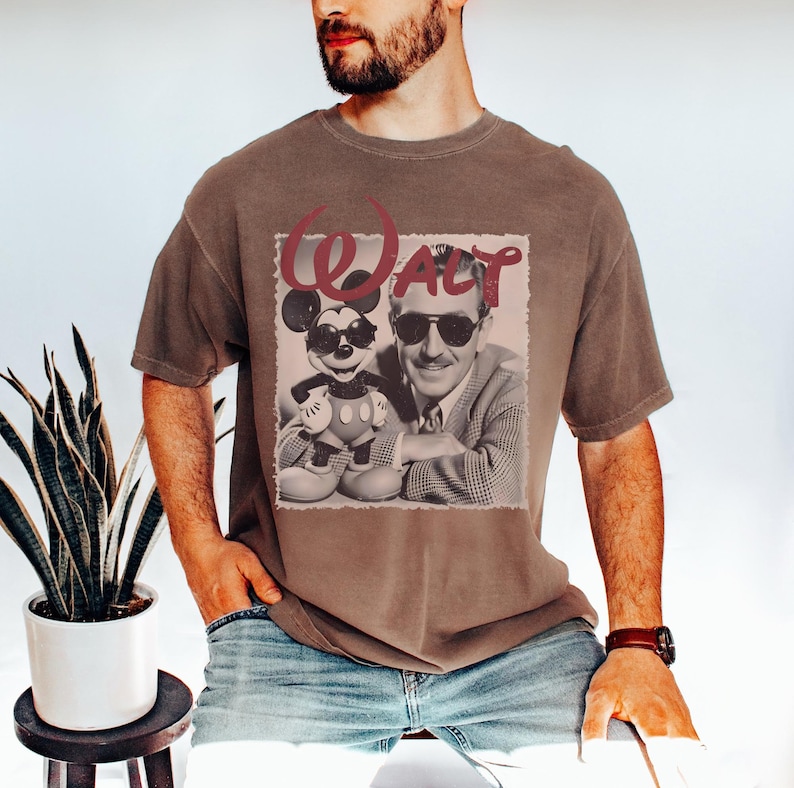 May include: A brown short-sleeved t-shirt featuring a graphic print of a black and white photograph of Walt Disney and Mickey Mouse, both wearing sunglasses. The word "Walt" is printed in large red script lettering above the central image.