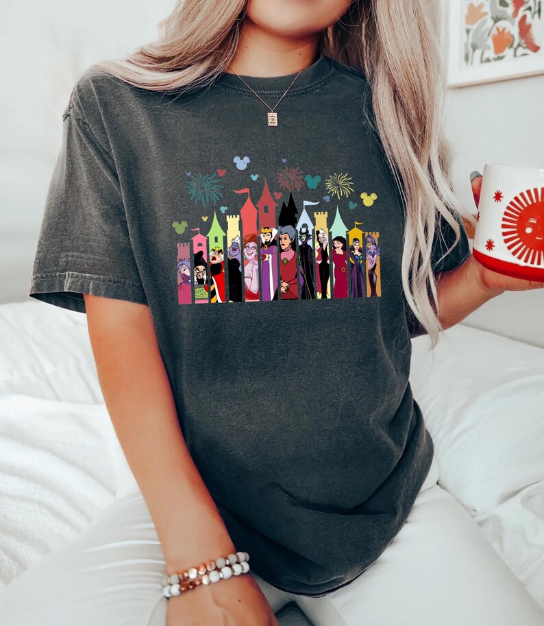 Puede incluir: Camiseta gris oscuro con un gr&aacute;fico colorido de villanos de dibujos animados frente a un horizonte de castillo. El gr&aacute;fico incluye fuegos artificiales y siluetas de Mickey Mouse. La camiseta tiene mangas cortas y un corte holgado.