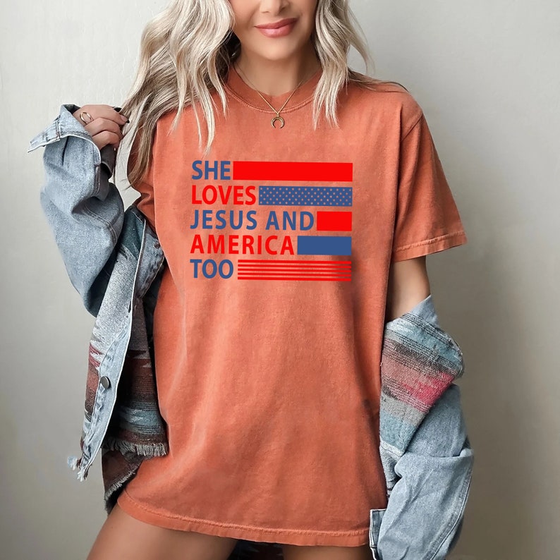 Może przedstawiać: Koszulka w kolorze wypalanej pomarańczy z napisem "She loves Jesus and America too" w kolorze czerwonym, białym i niebieskim. Tekst jest nałożony na grafikę z czerwonymi i niebieskimi paskami z białymi gwiazdami.