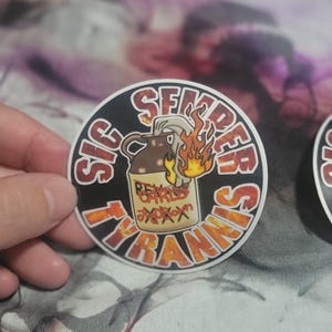 May include: A black and white round sticker with the text "Sic Semper Tyrannis" in a circular design. The sticker features a cartoon image of a lit Molotov cocktail with the text "Real Carls Jr. Joboxx" on the side.