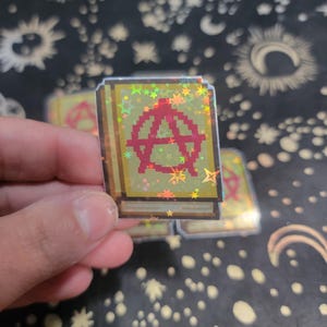 May include: A holographic sticker in the shape of a book with a pixelated design. The cover features a red anarchy symbol. The sticker has a gold and green background with star-shaped holographic accents.