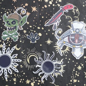 May include: A collection of stickers with a dark background and gold stars and moons. The stickers include a green goblin head with a hat and crossed bones, a bloody knife, a mechanical eye, a hand print, and sun designs.