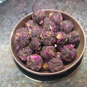 May include: A bowl filled with small, dark brown and purple tea balls. The tea balls are made with dried rose petals and other herbs.