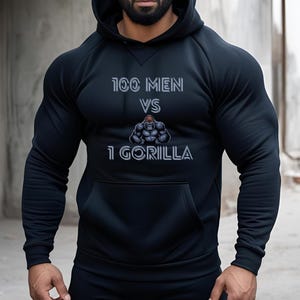 Puede incluir: Sudadera con capucha negra con el texto "100 MEN VS 1 GORILLA" en gris. El gráfico presenta una ilustración de gorila. La sudadera tiene un bolsillo delantero y una capucha con cordón.