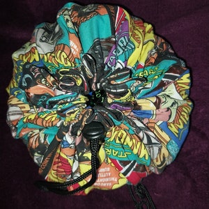 May include: A colourful fabric dice bag with a drawstring closure. The bag is made from a variety of comic book covers, including Star Wars, Wonder Woman, and other superhero characters. The bag is tied with a black drawstring and has a black button.