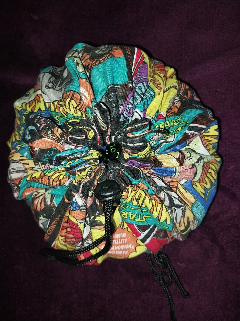 May include: A colourful fabric dice bag with a drawstring closure. The bag is made from a variety of comic book covers, including Star Wars, Wonder Woman, and other superhero characters. The bag is tied with a black drawstring and has a black button.