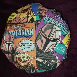May include: A round fabric cushion with a colourful patchwork design featuring characters and scenes from the Star Wars franchise, including The Mandalorian, Baby Yoda, and the text "Star Wars The Mandalorian".