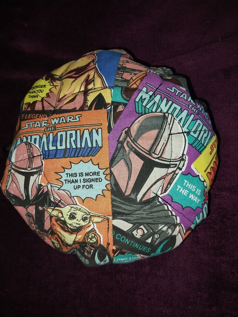 May include: A round fabric cushion with a colourful patchwork design featuring characters and scenes from the Star Wars franchise, including The Mandalorian, Baby Yoda, and the text "Star Wars The Mandalorian".