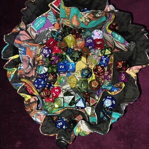 May include: A black fabric dice bag with a comic book print design. The bag is filled with a variety of colourful dice, including clear, red, blue, green, and gold dice.