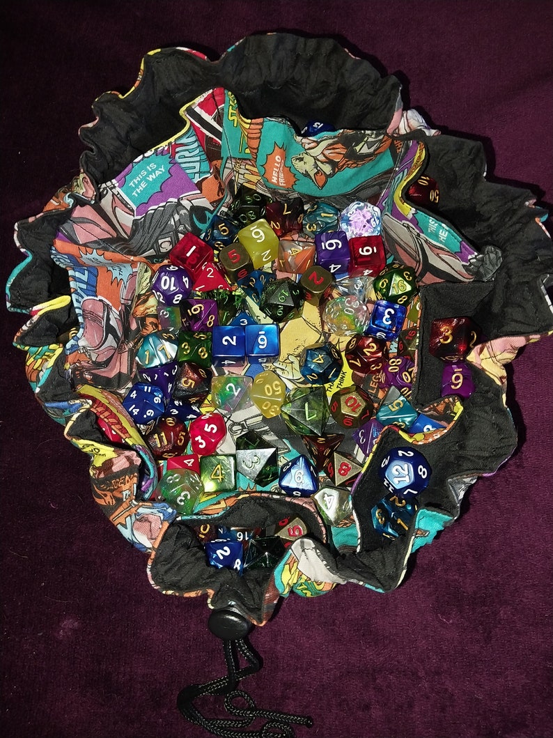 May include: A black fabric dice bag with a comic book print design. The bag is filled with a variety of colourful dice, including clear, red, blue, green, and gold dice.