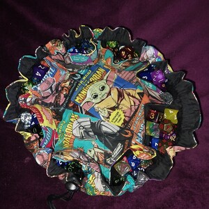 May include: A black fabric dice bag with a drawstring closure. The bag is decorated with a colourful comic book pattern featuring characters from Star Wars. The bag is filled with various coloured dice.